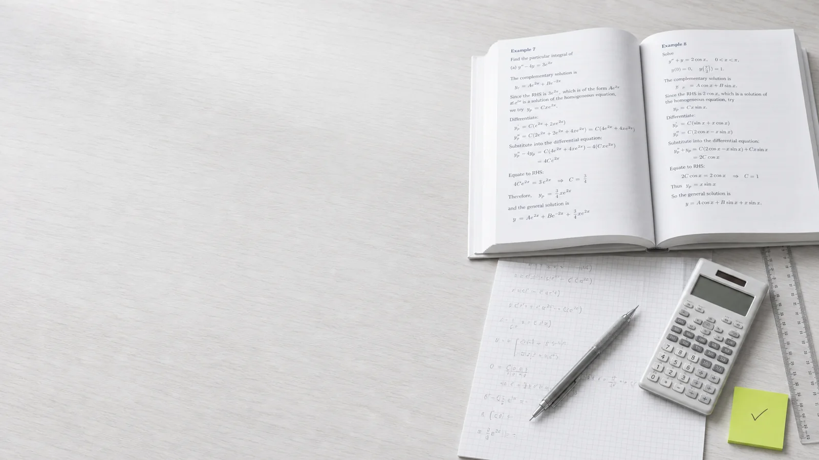Editorial still-life of an open A-Level Maths textbook with worked pencil solutions on graph paper